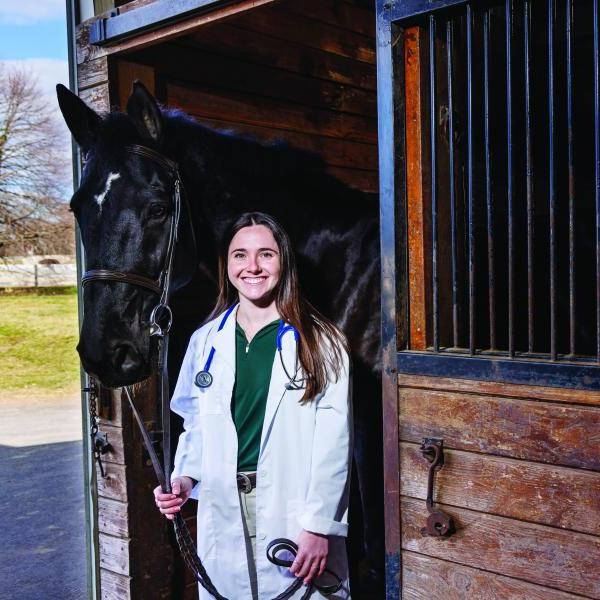 尼娜Dellarte, a class of 2021 graduate is in a white coat next to a black horse at the equestrian center.